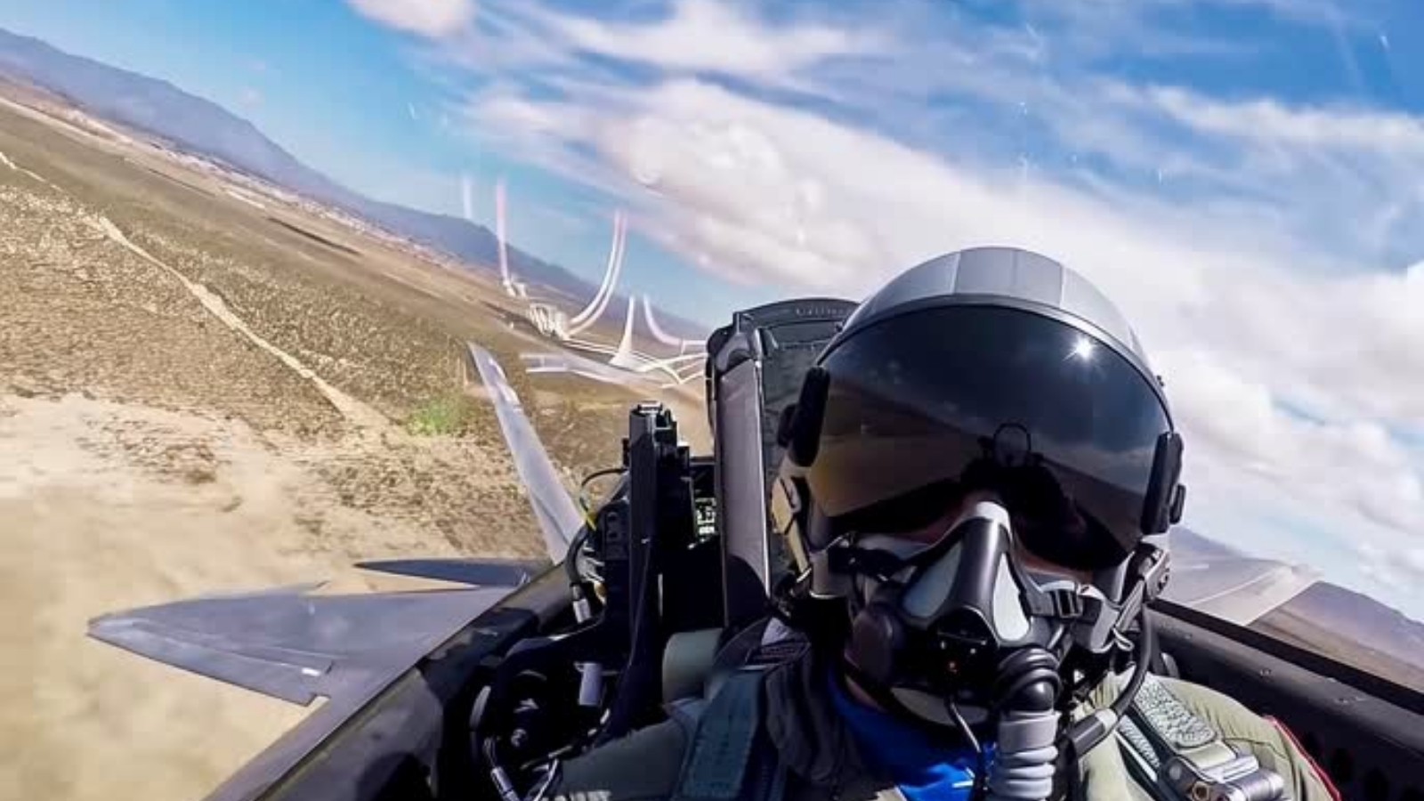 A fighter pilot in a helmet and oxygen mask sits in the cockpit of a jet flying low over desert terrain.