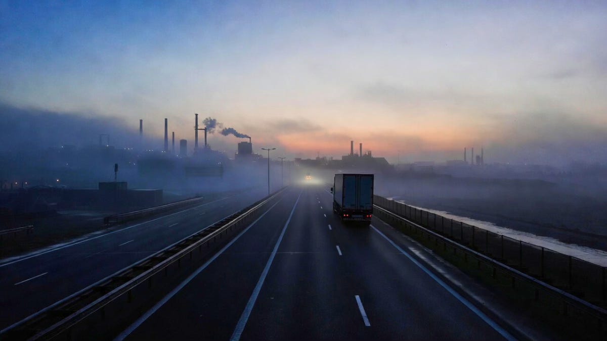 A truck drives down an empty highway at dawn toward a row of factory smokestacks in the haze.